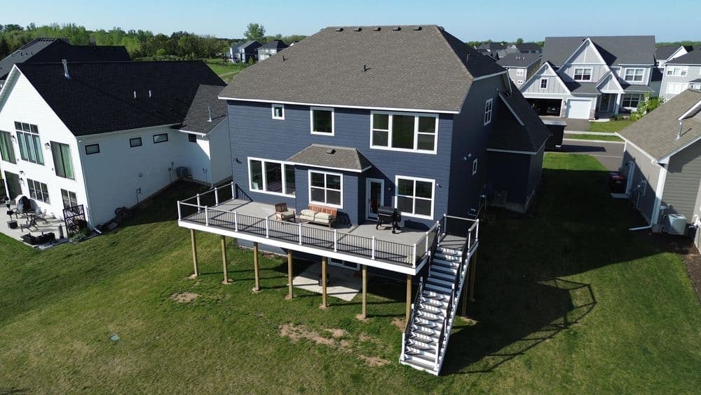Modern two-story house with a deck, situated in a suburban neighborhood. Green lawn and blue skies.